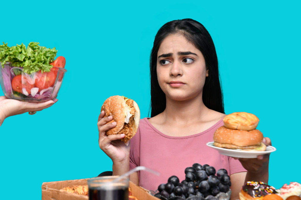 202606-blog-alimentos-inflaman-1 » Pura Prote Young woman looks unsure, holding a hamburger and a plate of donuts while a salad bowl is offered from the left.