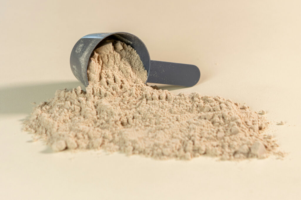 Grey measuring scoop tipped over, spilling beige powder onto a light surface, creating a small mound and scattered grains.